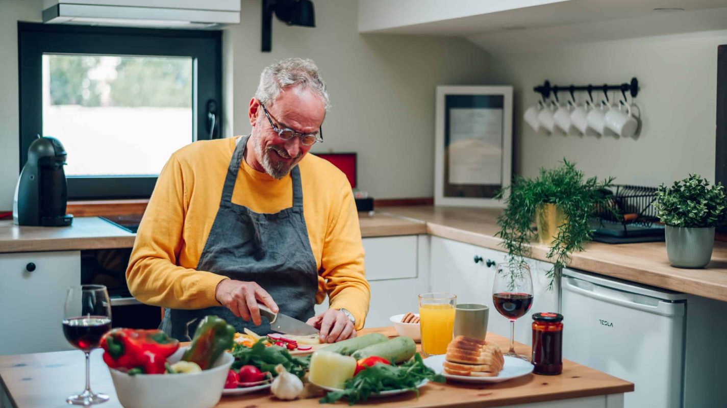 Elderly man preparing dinner for his family while cutting vegetables in the kitchen and cooking food meal at home. Portrait of a senior man in a modern kitchen. Copy space.